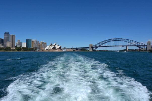 Toàn cảnh Sydney Harbour với Nhà hát Opera Sydney và cầu cảng Harbour Bridge nhìn từ phía sau một chiếc phà trên mặt nước xanh vào ban ngày.